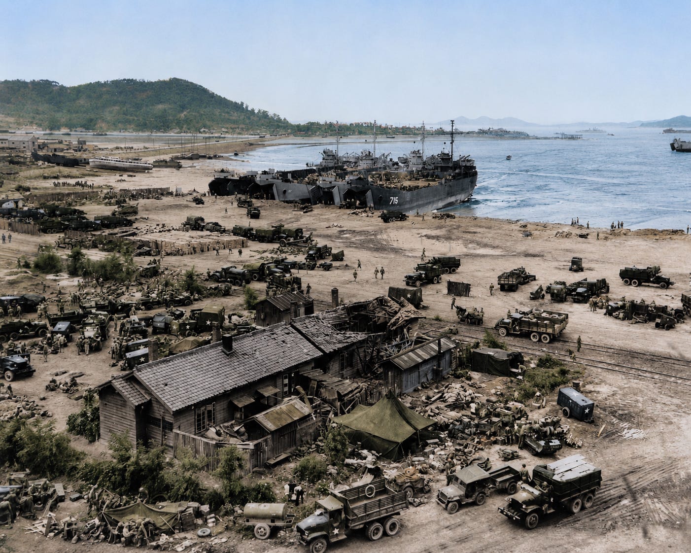 Military vehicles and equipment are assembled near a coastal landing site, with several large naval ships docked on the shore and hills in the background.