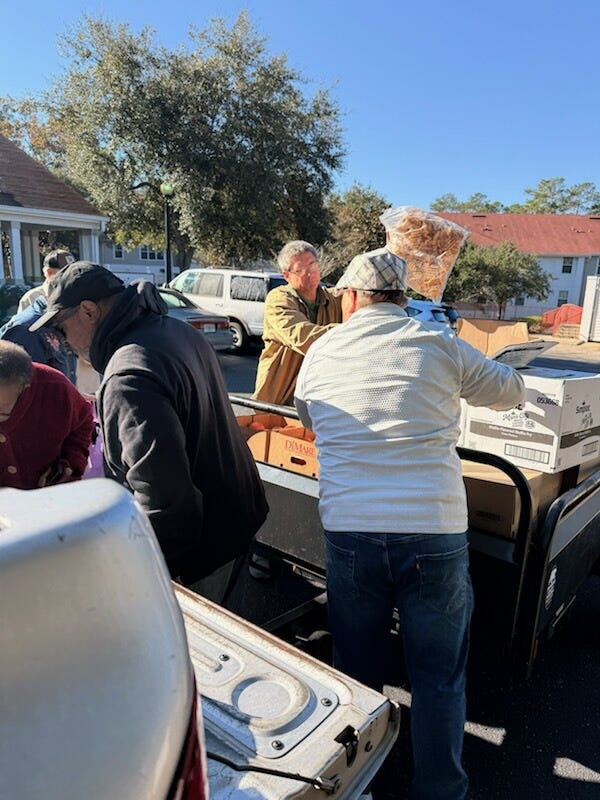 A group of adults work together outdoors in a sunny residential parking lot, transferring cardboard boxes and a large clear bag of food from a pickup truck to a black Club Car utility cart. Parked cars, trees, and houses with red roofs are in the background. A group of adults work together outdoors in a sunny residential parking lot, transferring cardboard boxes and a large clear bag of food from a pickup truck to a black Club Car utility cart. Parked cars, trees, and houses with red roofs are in the background.