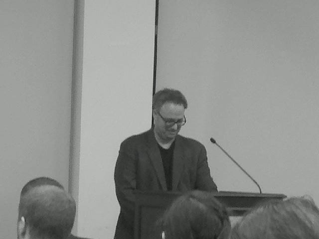Photo of me, Daniel Nester, standing at a podium. I am looking down at my notes and a long microphone is front of my face. It's 2013 and my hair is not yet gray.