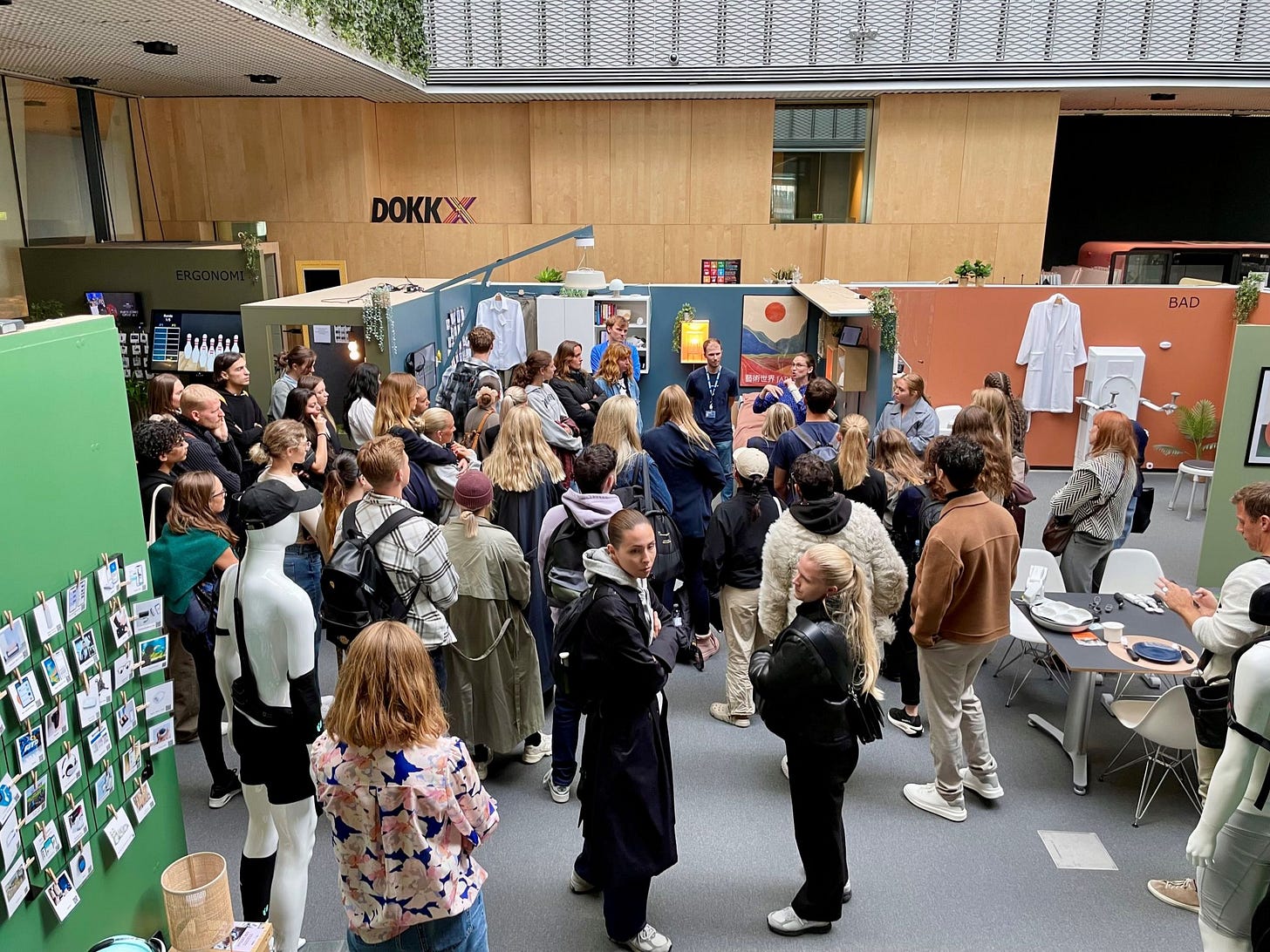 An exhibition or trade show at DOKKX featuring mock-up displays of different rooms. A large crowd of attendees is gathered around colorful partitioned displays that showcase different living spaces, including a bathroom area labeled "BAD" with white fixtures. The space has wood-paneled walls with the DOKKX logo displayed prominently. Various display areas feature different themes, including one labeled "ERGONOMI." Several mannequins are positioned throughout the space, and information boards with small images are visible on a green partition. Attendees appear to be listening to a presentation or guided tour.
