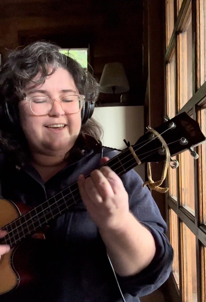 Two images: 1, a close-up of a tiny, colorful paper laundry basket, a little booklet that says "Laundry Divination How-To Guide", and a spread of brightly colored paper-doll sized clothes, 2 :Devan and her ukulele by a window in a dark-paneled room, smiling as she plays