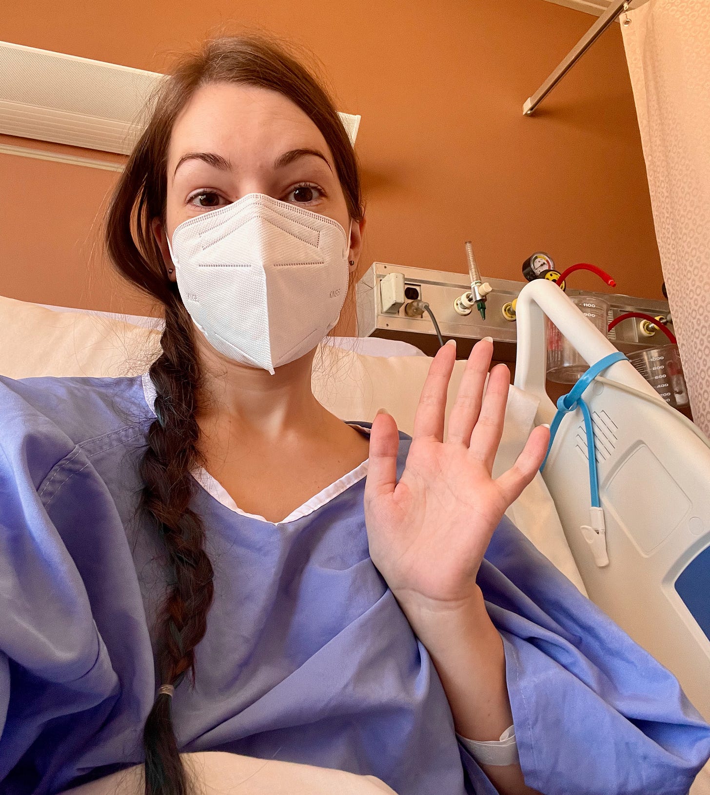 A light-skinned woman in a hospital bed wearing a blue hospital gown, waving to the camera.