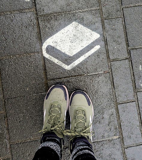 Three images. The first is myself in a yellow knit hat in a forest facing down toward the camera with trees and sunlight on Walden Pond. The middle image is The Bookhive logo, which is a stack of books in the shape of a hive with the words underneath. The third image is looking down towards my green shoes with a white book painted on a paved walkway.