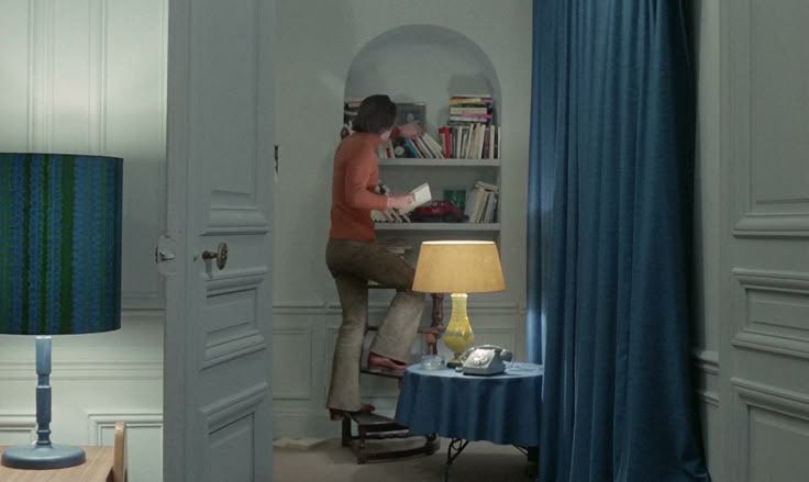 This may contain: a woman standing in the doorway of a room with a lamp and bookshelf