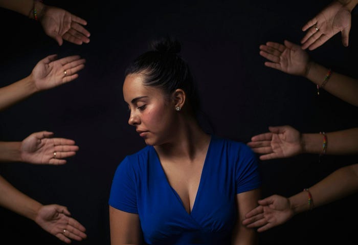 A woman in a blue dress is seen in a contemplative pose with her head turned to the side, surrounded by multiple outstretched hands against a dark background. A woman in a blue dress is seen in a contemplative pose with her head turned to the side, surrounded by multiple outstretched hands against a dark background.