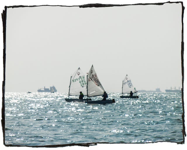 Three small sailboats with single passengers out at sea. There are large fishing boats on the horizon.