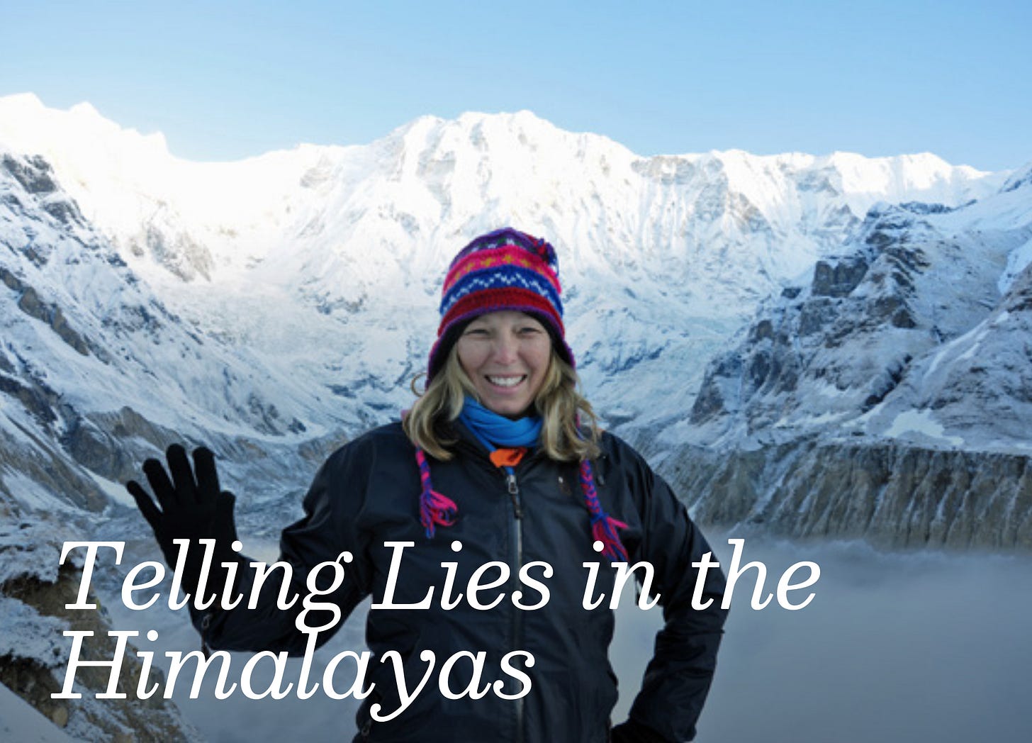 White woman wearing a colorful knit hat and black hiking clothes smiles while posing in front of snow-covered peaks.  
