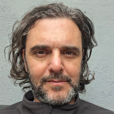 Homem pardo de cabelo comprido, até o ombro e barba grisalha, usa um casaco marrom de gola alta sem estampa e sorri levemente para a câmera de costas para uma parede esverdeada. Homem pardo de cabelo comprido, até o ombro e barba grisalha, usa um casaco marrom de gola alta sem estampa e sorri levemente para a câmera de costas para uma parede esverdeada.