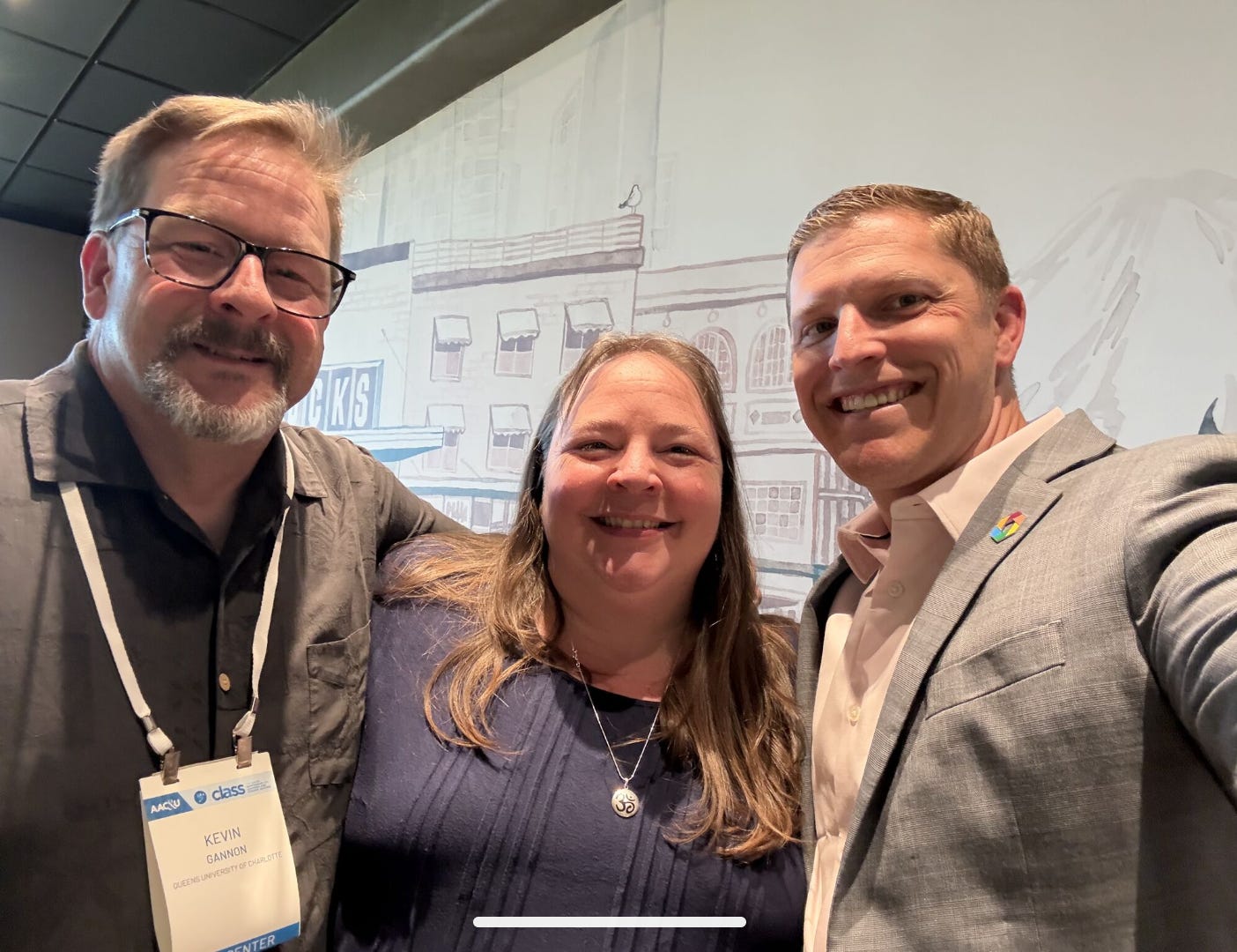 Three adults stand with arms linked at a conference center Starbucks. On the left is Kevin Gannon, a tall white man with a goatee and glasses, wearing a black button-down shirt and a nametag. In the middle is Liz Norell, an average height white woman with long hair and a round face, wearing a navy blue shirt and a silver necklace with the sanskrit symbol for ohm. On the right is Kevin McClure, a white man wearing a gray suit jacket, a white button down shirt, and a lapel pin with the logo from his book, The Caring University.