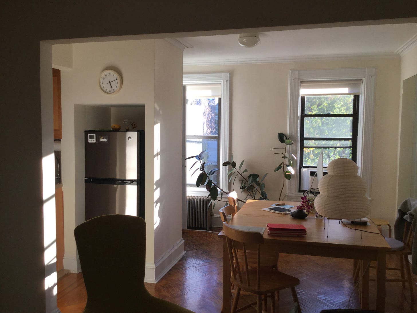 A view into an apartment. You can see a refrigerator in a nook, a table with a desktop computer and a paper lamp, and a lush plant against the back wall.