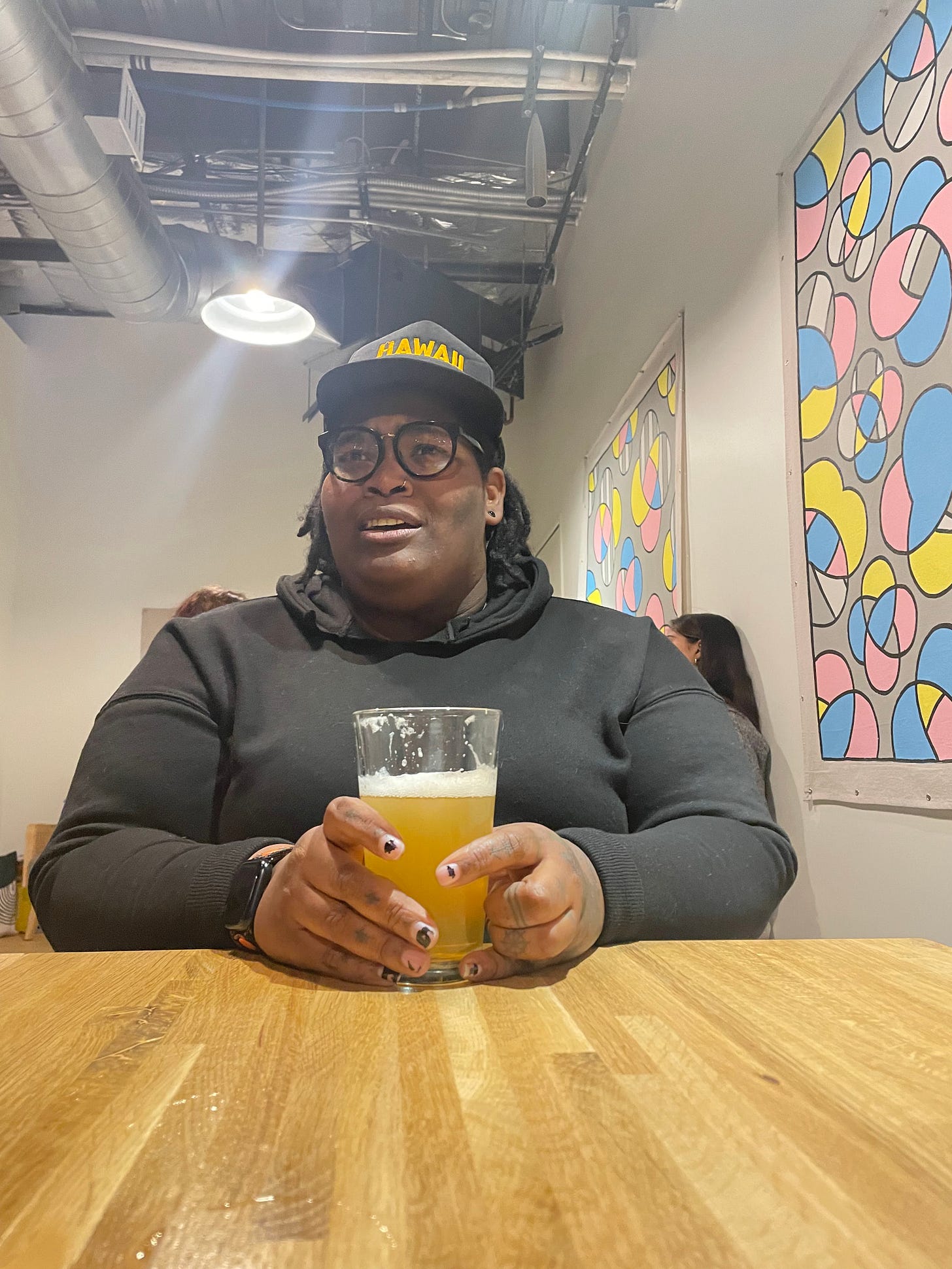 A black nonbinary person with locs, a black trucker hat and black hoodie holding a class of non-alcoholic beer at a table with pastel art behind them.