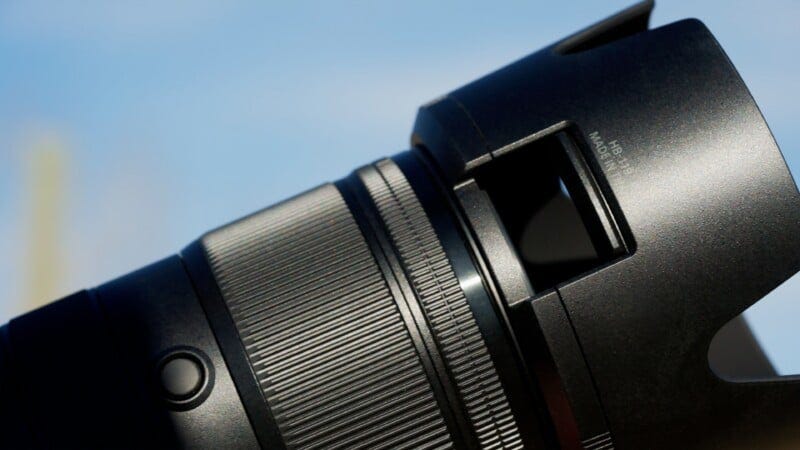Close-up of a black camera lens set against a clear blue sky, showing detailed texture, ridges, and buttons on the lens barrel. Close-up of a black camera lens set against a clear blue sky, showing detailed texture, ridges, and buttons on the lens barrel.