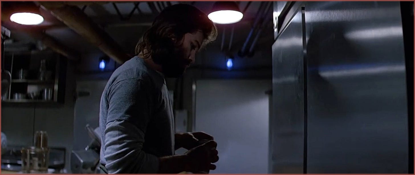 Interior shot from The Thing (1982): MacReady stands in profile in a dimly lit room, two china hat lanterns visible on the ceiling above him casting downward light, exposed pipes and ductwork illuminated overhead. Two blue practical lights glow in the background. Dean Cundey's dual lighting system — actors below, architecture above — visible in a single frame.