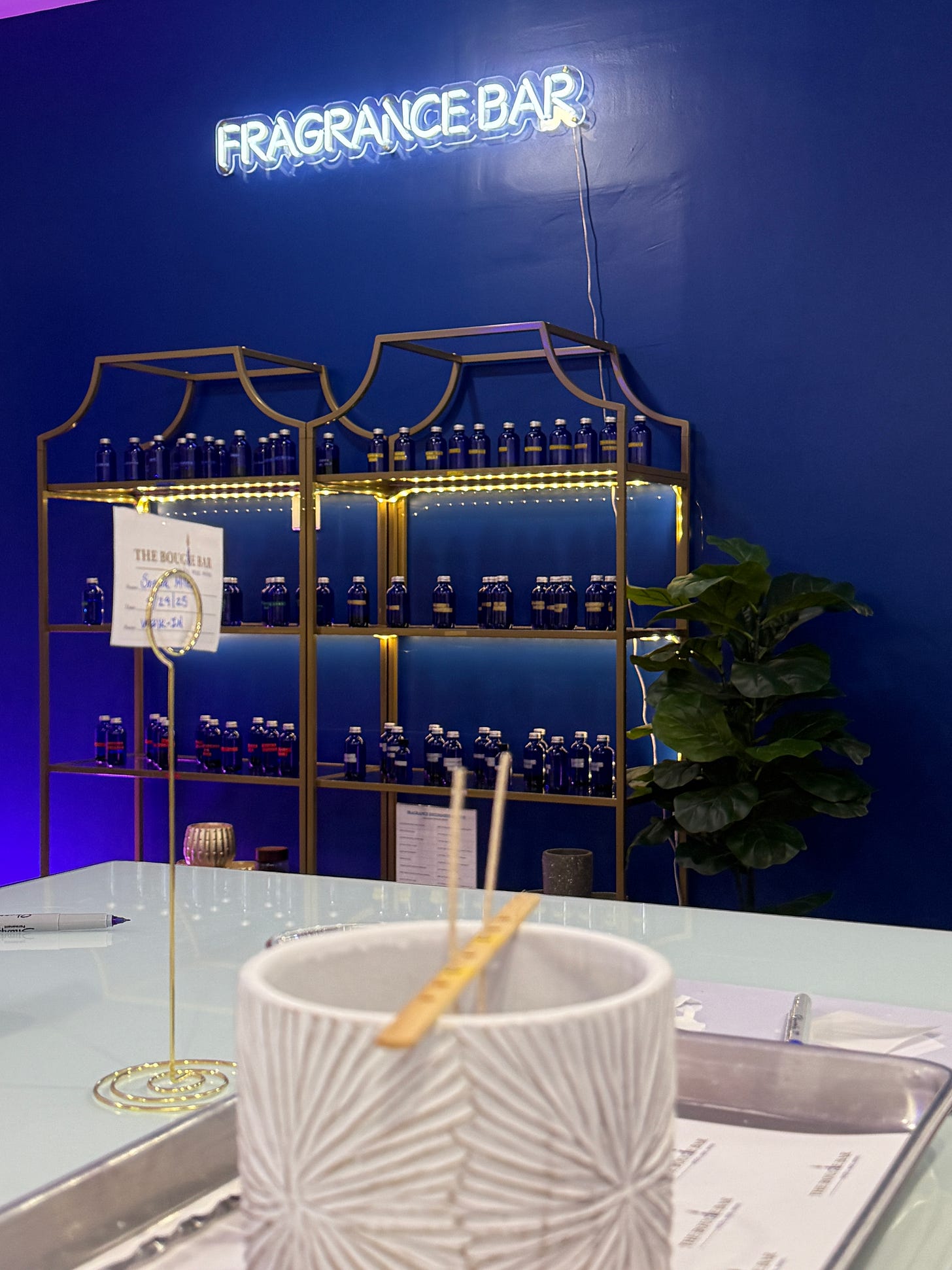 Wall display of blue glass fragrance bottles on gold shelving at The Bougie Bar, with neon 'Fragrance Bar' sign above Wall display of blue glass fragrance bottles on gold shelving at The Bougie Bar, with neon 'Fragrance Bar' sign above