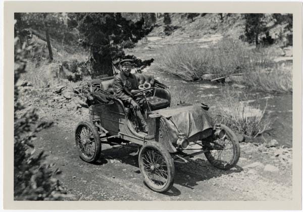 Old black and white photo of Alexander Winton driving his Winton autmotobile on a gravel track alongside a river. The automobile consists of four tall and narrow wire spoked wheels and an open air seating area with a thickly-cushioned leather bench seat and a slender steering column extending up from the floor to a nearly vertical steering wheel. Winton is seated in the driver's seat (on the right). He is wearing what looks to be a leather or waxed cotton jacket, dark leggings/trousers, and a driving cap. He is looking at the camera. He has a thick mustache.