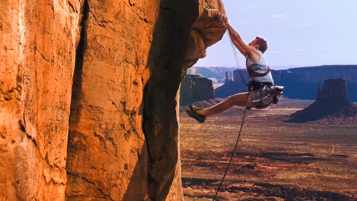A climber clings in a pike position to a steep red rock wall in Monument Valley, hoisting himself up an outcropping, with a sweeping vista of mesas in the background.