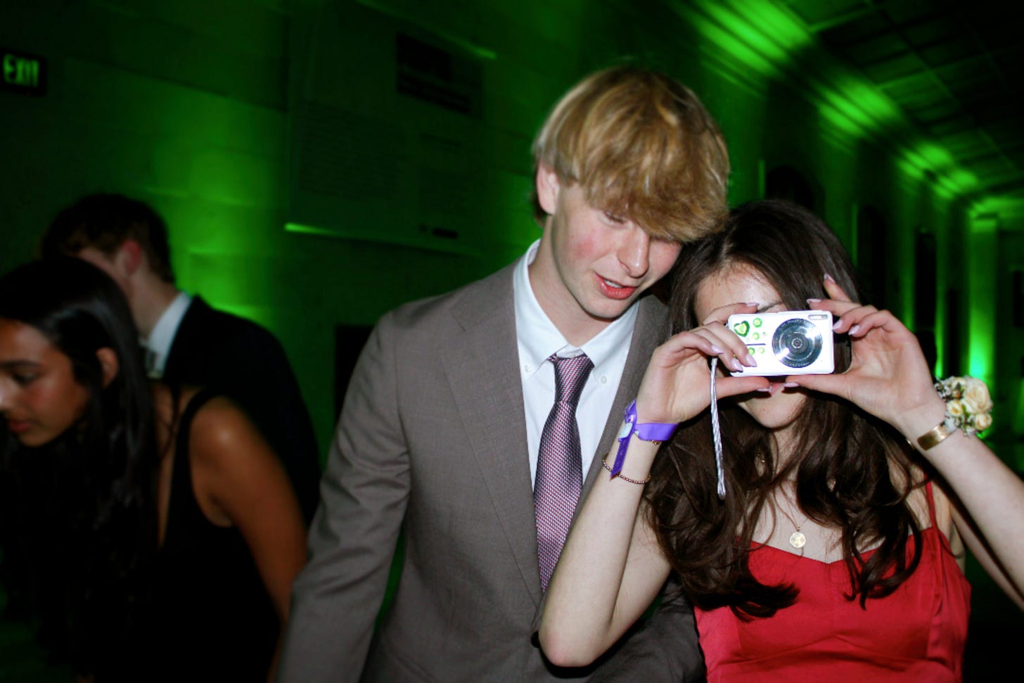 A young woman in a red dress holds a camera to her face, standing beside a young man in a suit and tie. The background is illuminated with green lighting.