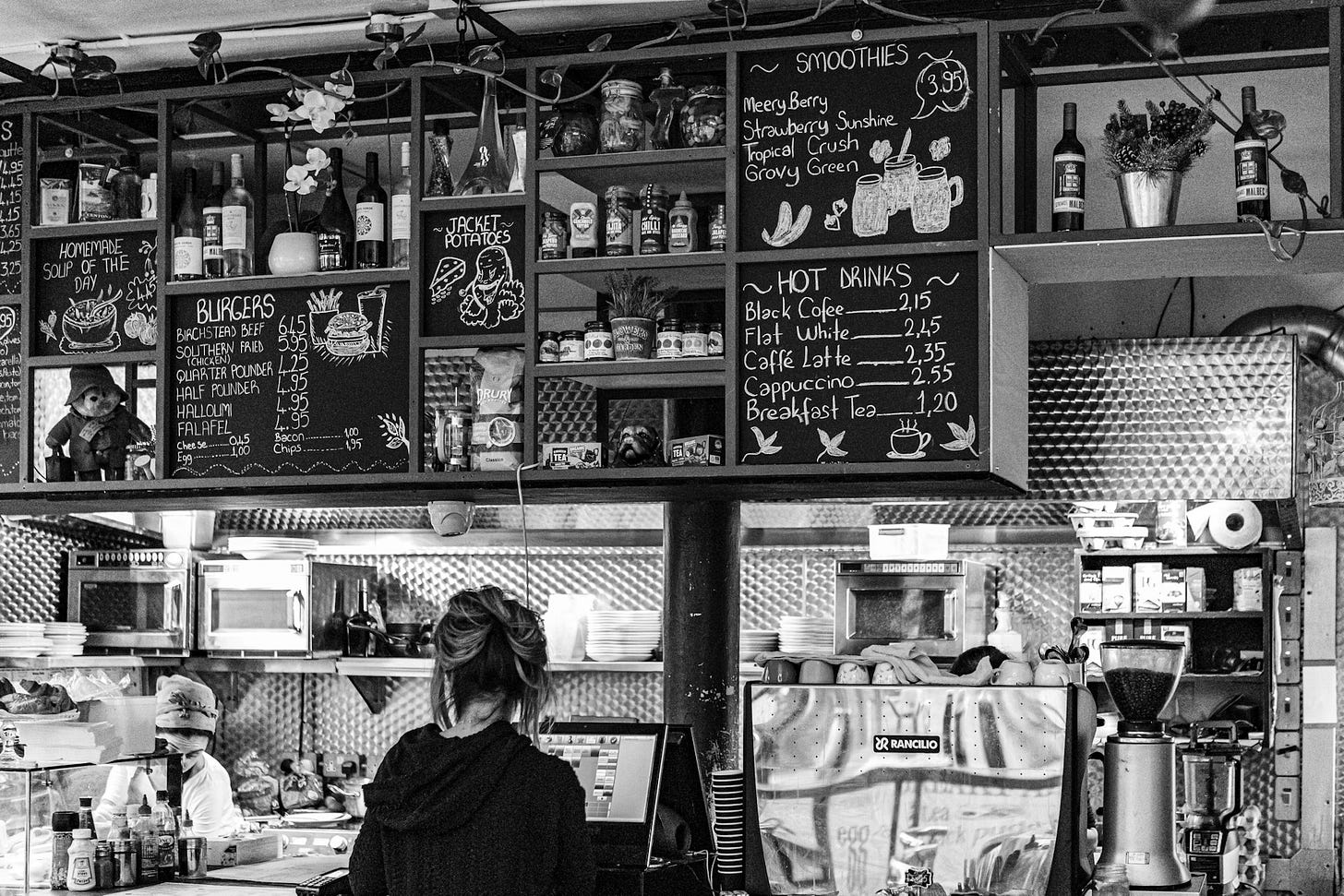 A black and white photo of a small London café interior showing a staff member at the counter. Chalkboard menus display the prices for various items including burgers, smoothies, and hot drinks. The kitchen area and storage shelves with bottles and supplies are visible in the background.