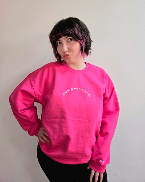 Modeling merch: A hot pink sweatshirt with white embroidered text on the front chest that says "gluten gives me the ick" and "GFGC" on the left sleeve near the wrist. A vintage wash denim baseball cap with an embroidered white pixelated chat bubble and black "GFGC" inside. A black sweatshirt with a green embroidered design on the front chest that says "GAME OVER, YOU ATE GLUTEN AND DIED" with a pixelated ox and wagon and "GFGC" on the left sleeve near the wrist.