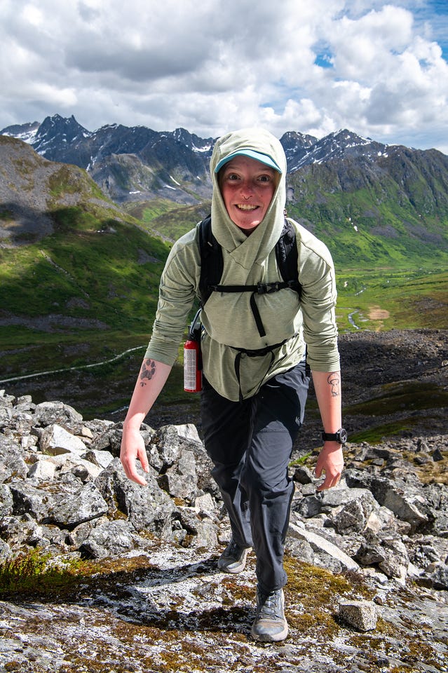 A smiling hiker in a pale green sun hoody climbs directly toward the camera across lichen-covered rock, a black pack on her shoulders and a canister of bear spray clipped at her chest, small tattoos visible on both forearms. Behind and below her, a broad green valley floor winds between dark, snow-streaked ridgelines under a sky of broken cloud. A smiling hiker in a pale green sun hoody climbs directly toward the camera across lichen-covered rock, a black pack on her shoulders and a canister of bear spray clipped at her chest, small tattoos visible on both forearms. Behind and below her, a broad green valley floor winds between dark, snow-streaked ridgelines under a sky of broken cloud.