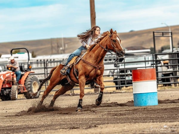 A rider skillfully navigates a horse around barrels during a rodeo event, with a tractor and spectators in the background.