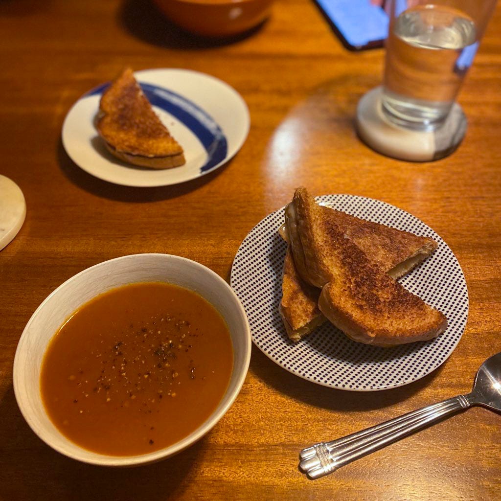 A small white bowl of tomato soup next to a browned grilled cheese sandwich, sliced in half diagonally, on a blue and white plate. In the background is another sandwich half on a different plate, and a glass of water on a coaster.