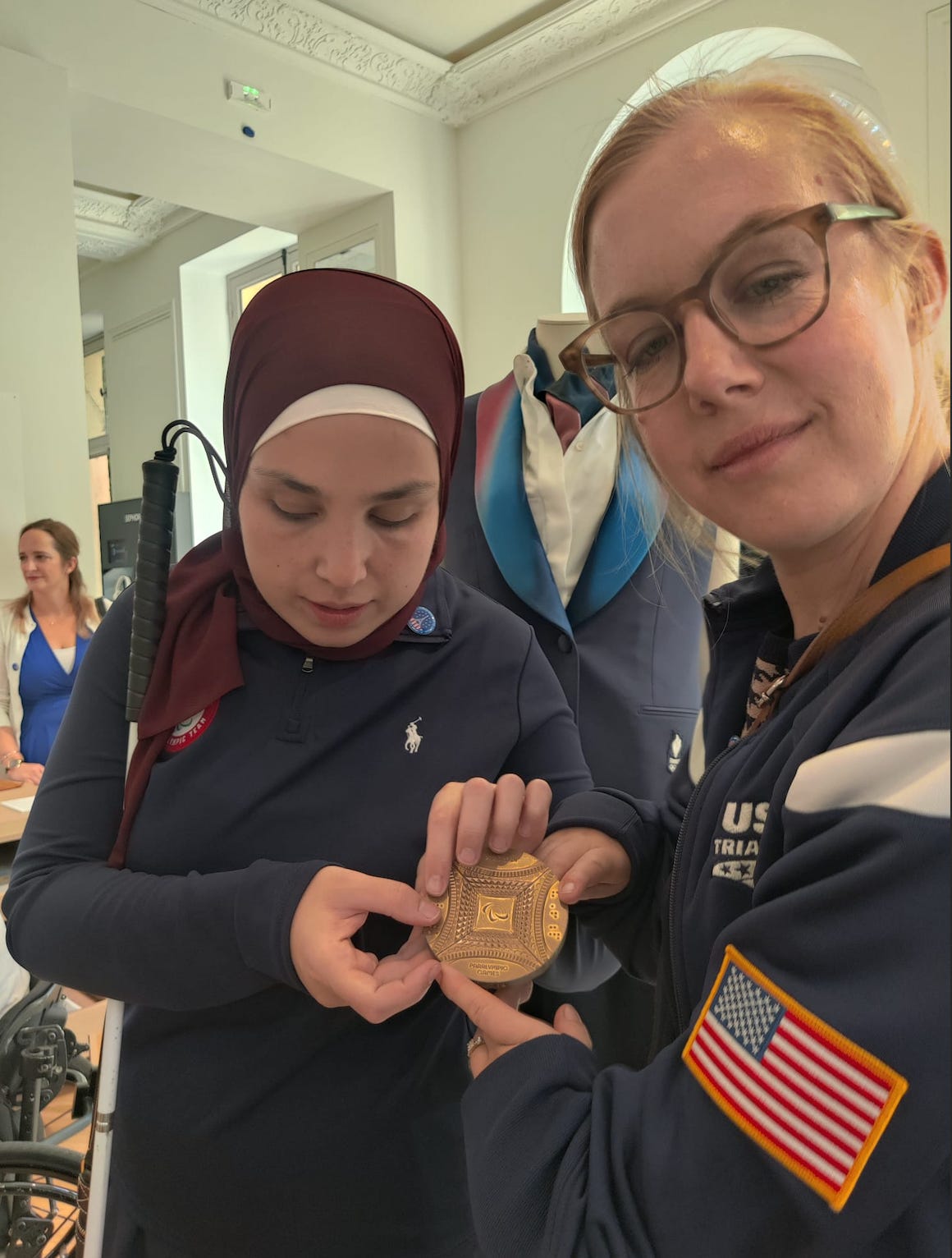Sara stands with a member of Team USA on her right. She touches the Olympian’s bronze medal, which includes brail. Sara wears a navy team USA quarter-zip sweater and a burgundy red hijab. 