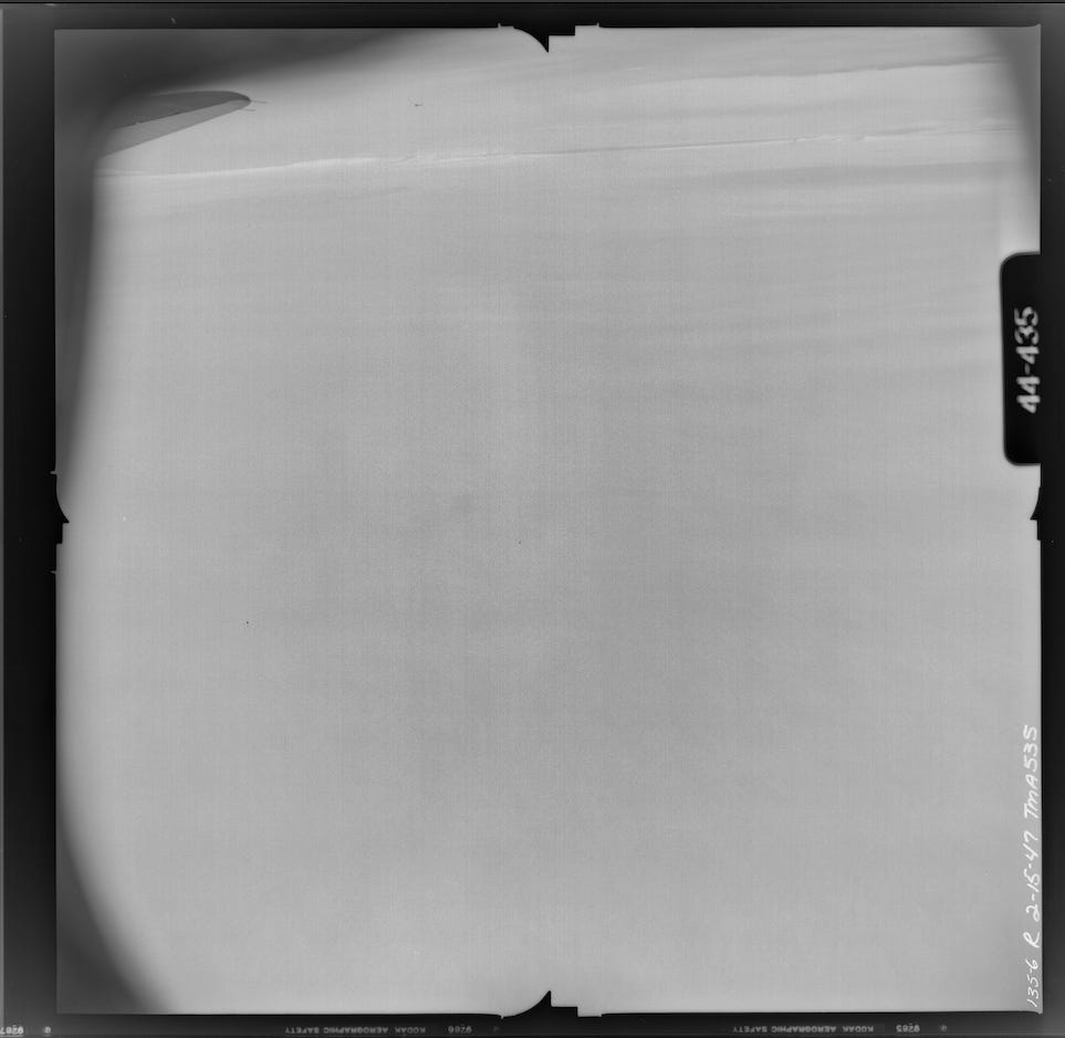 A greyscale photo of the surface of Thwaties Glacier. The tip of an airplane wing can be seen in the upper lefthand corner. No features are discernable on the surface except for the horizon. The surface is indistinct but the greys undulate slightly. The image is surrounded by a black frame.