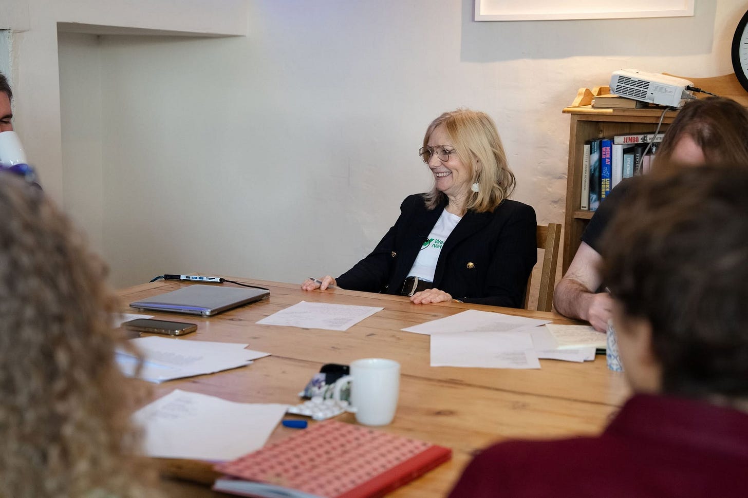 A photograph of Mayor Tracy Brabin sat around a table with numerous writers. The table is scattered with pages of poems, notebooks, mugs and pens. Tracy Brabin is holding a pen and smiling at someone out of frame. 