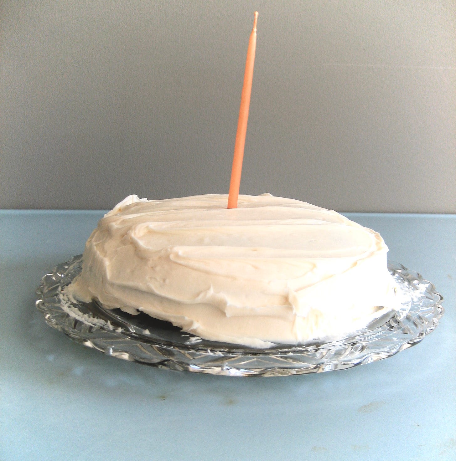 A photo of a circular cake with cream cheese frosting and one tall orange candle rising up from the center, plated on antique crystal that's sitting on a vintage blue metal table.