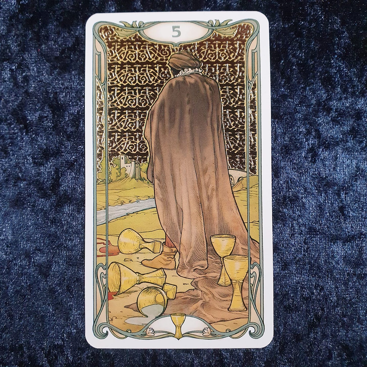 Five of Cups from the Golden Art Nouveau Tarot on blue velvet, showing a cloaked figure mourning spilled cups in a melancholy landscape, used in Tarot DMs with Emma van Straaten.