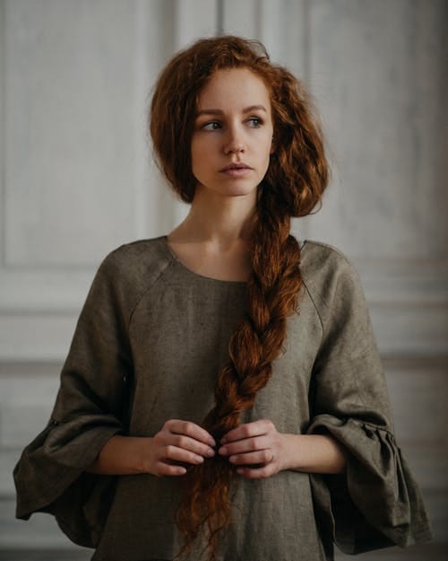 Free Serious female in green dress looking away while touching ginger hair in braid on blurred background Stock Photo