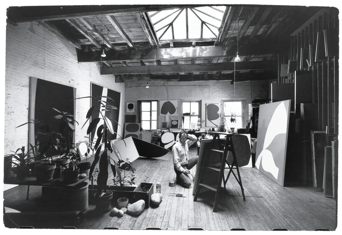 black and white photo of a loft-like space full of art and plants, with a man kneeling down in front of a canvas black and white photo of a loft-like space full of art and plants, with a man kneeling down in front of a canvas