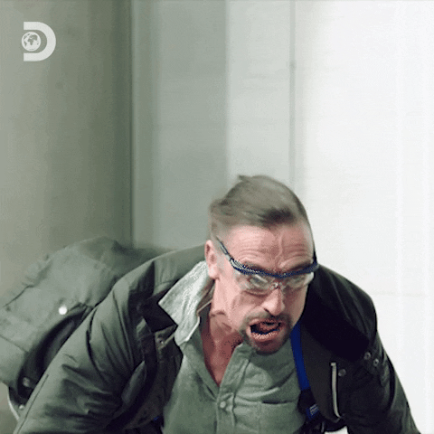 A man in what seems to be a strong wind tunnel, looking around in panic as he tries to remain upright.