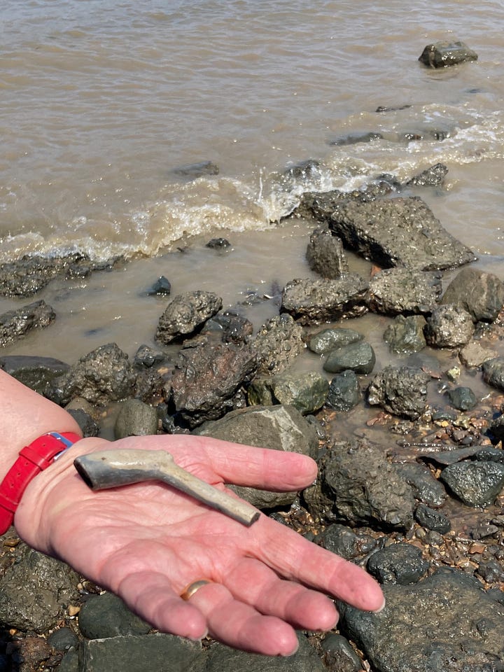 Man holding a canon ball, a shark's tooth and a typical mix of stones and pebbles on the banks of the river Thames