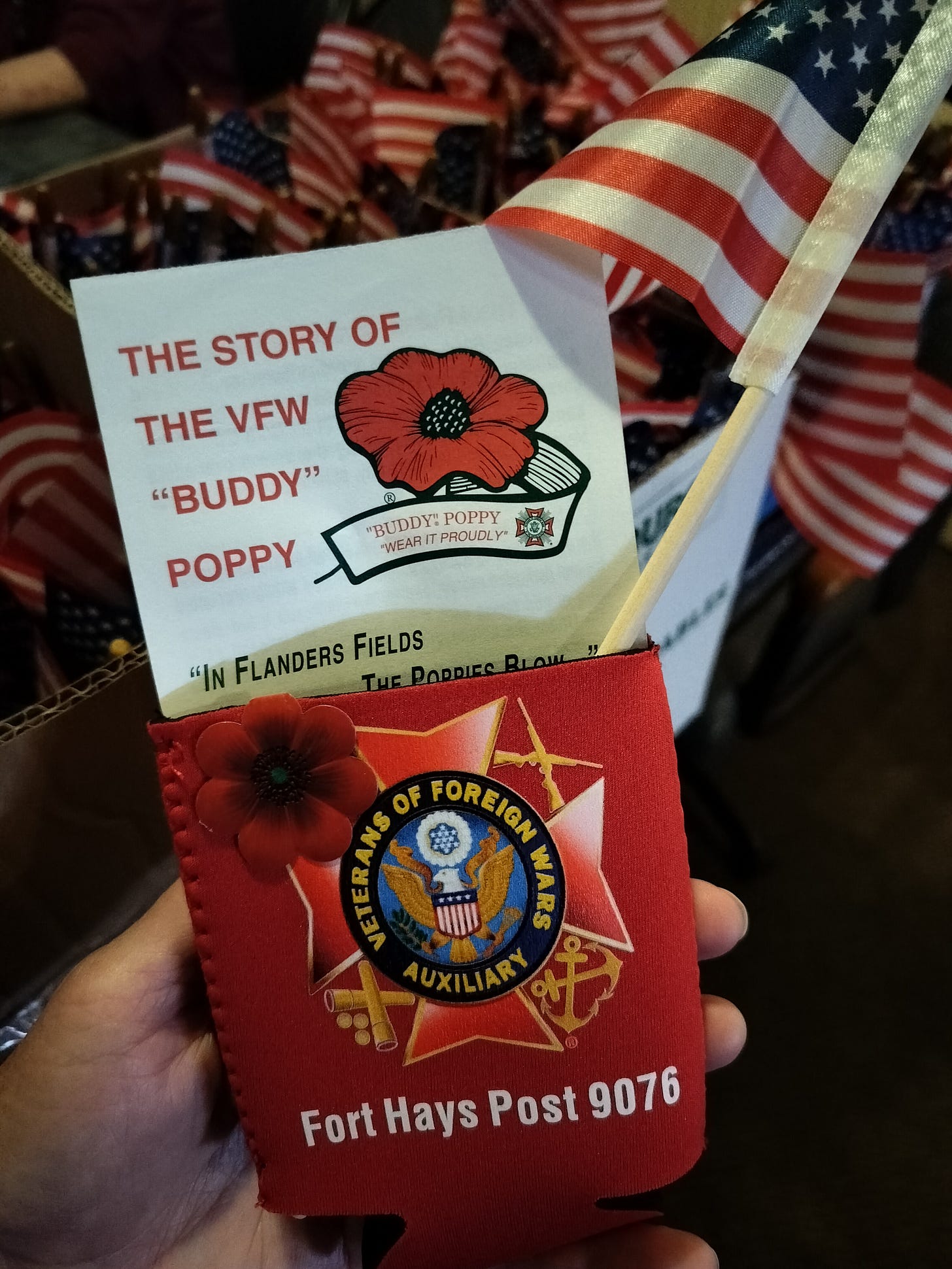 The author holding a red koozie with the VFW Auxiliary logo and a red poppy pin, with an American flag tucked inside.