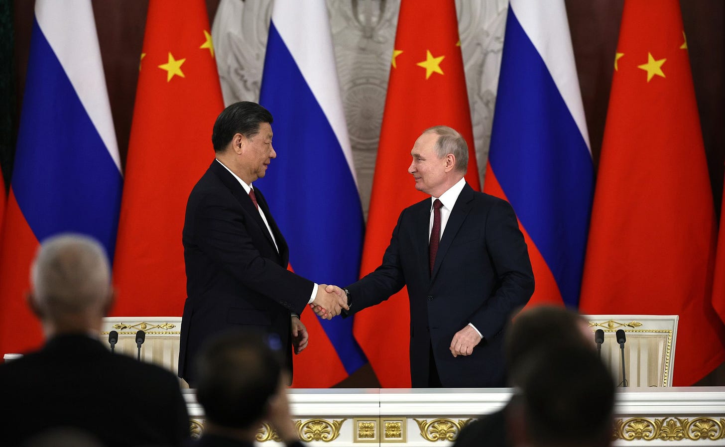 Fotografia em cores de Xi Jinping e Vladimir Putin em aperto de mãos em 2023. Ao fundo, bandeiras da China e da Rússia.
