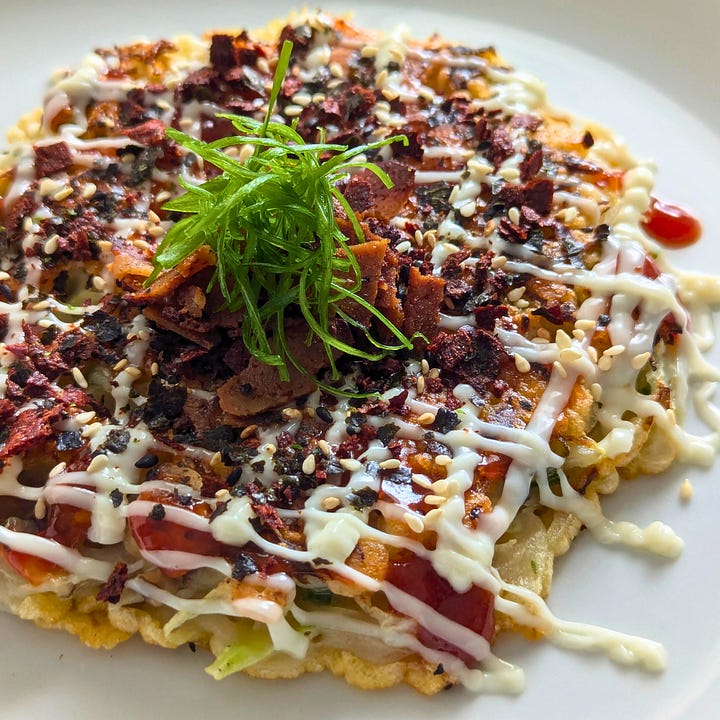 Two finished okonomiyaki pancakes, each topped with okonomiyaki sauce, vegan mayo, and different garnishes.