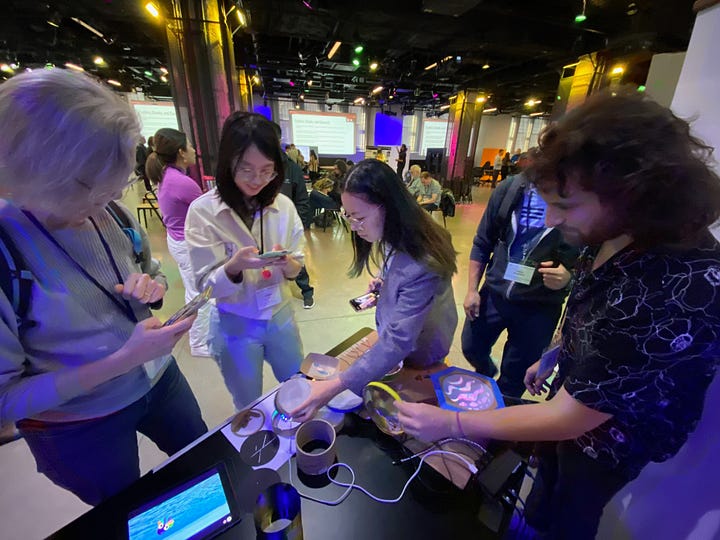 Photo 1: conference attendees gather around a table and interact with and take photos of materials including an iPad, cardboard tubes, LED lights, and physical materials. Photo 2: conference attendees gather around a table full of paper and craft materials and watch as a facilitator demonstrates placing a paper window cut-out on top of a phone screen.