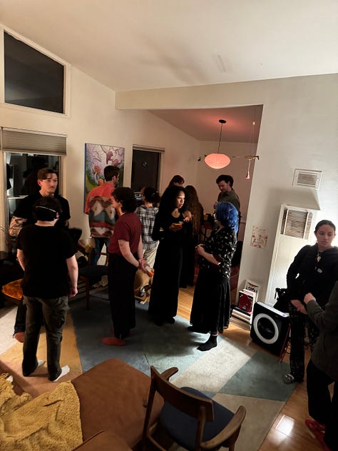 Photo 1: People gathering in a living room with vaulted ceilings. Photo 2: A painting and 3 drawings hung on a wall. Photo 3: A surrealist painting of amorphous creatures hanging from a tree. Photo 4: People gathered around the surrealist painting. Photo 5: A collage with cartoon images of a trail to the ocean and a butterfly, and bold text reading: traces of a path, losing sight of the sea. Photo 6: People and a red dog gathered around a man playing guitar. Photo 7: Ink and pastel drawings of two circles meeting. Photo 8: a pile of shoes at a door. Photo 9: Silhouettes of people through tall windows from outside at night.