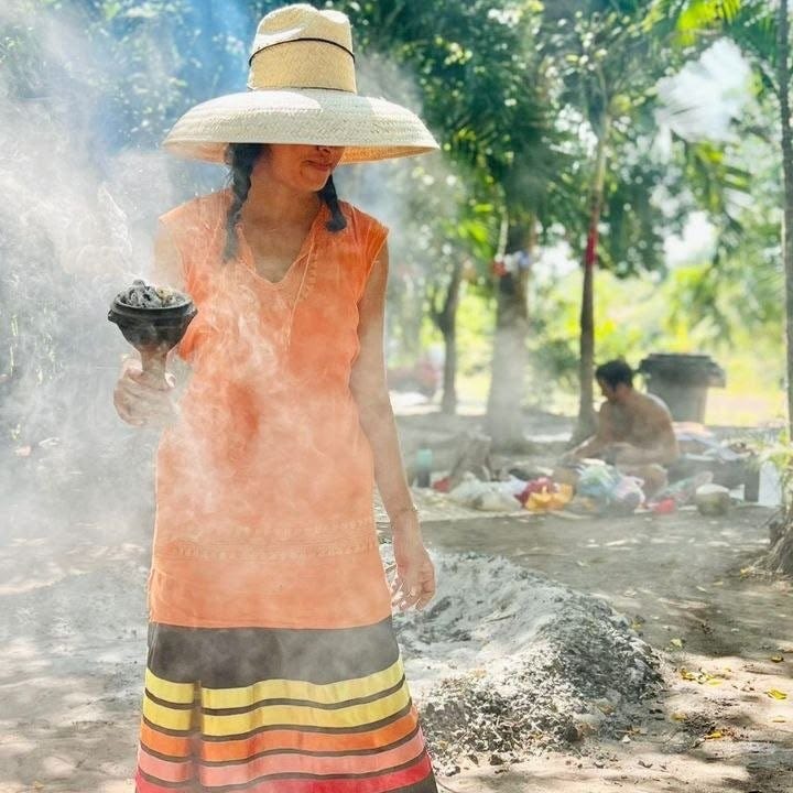 A ceremony leader in a woven palm hat and embroidered tunic raises a smoking copal censer in a sunlit clearing