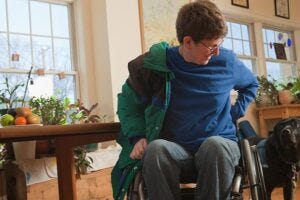 Woman with multiple sclerosis in a wheelchair putting on her coat with service dog watching her