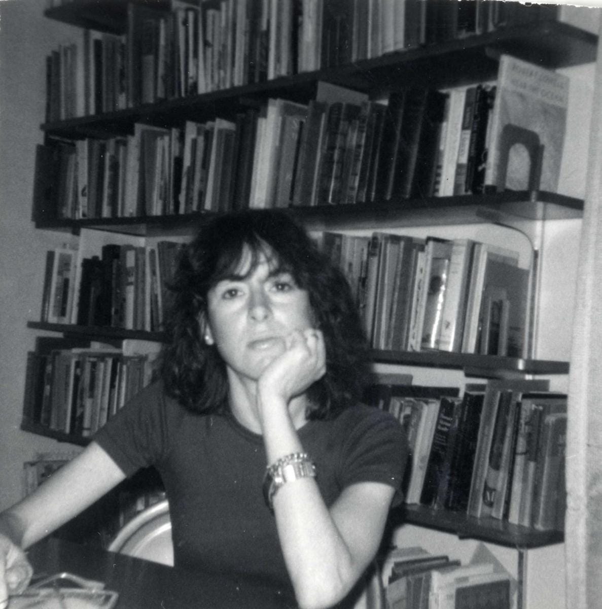 Louise Glück está sentada à mesa com a mão sob o queixo em frente a uma estante de livros