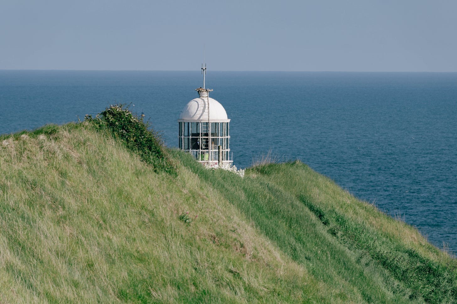 A white lighthouse dome peeks above green cliffs overlooking the blue Irish Sea at Howth Head A white lighthouse dome peeks above green cliffs overlooking the blue Irish Sea at Howth Head