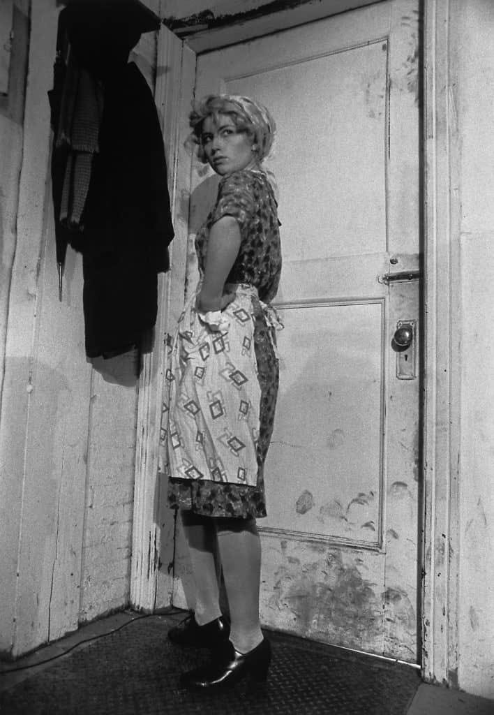 Foto in bianco e nero di una ragazza con un vestito a fantasia, con un grembiule. Ha anche una fascia tra i capelli. La foto è intera, guarda dietro di sè con sguardo sospetto, ha una mano sul fianco.