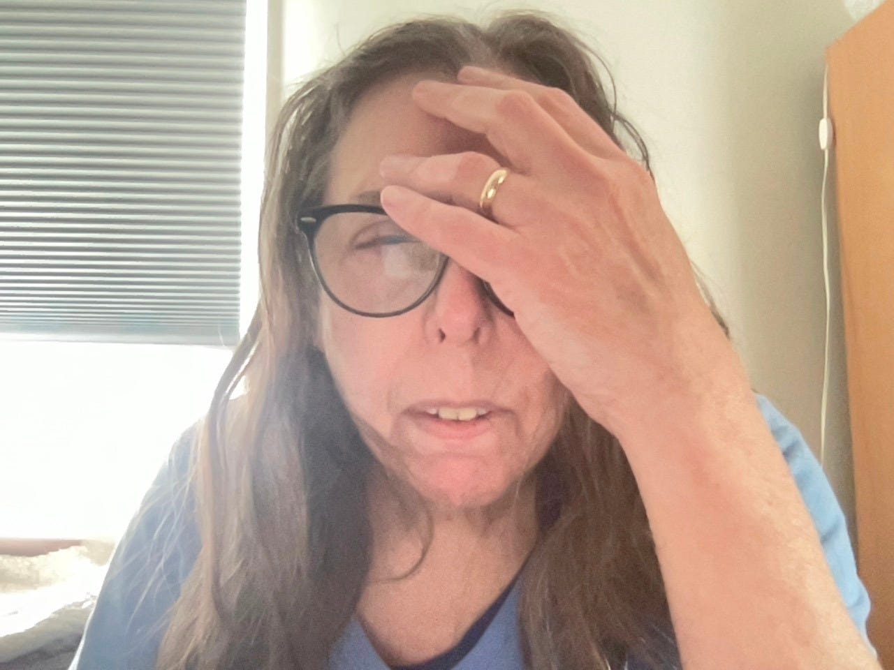 Woman with long scraggly brown hair, wearing a blue shirt, dark-rimmed glasses, frowning in pain as she holds the left side of her face because of migraine.