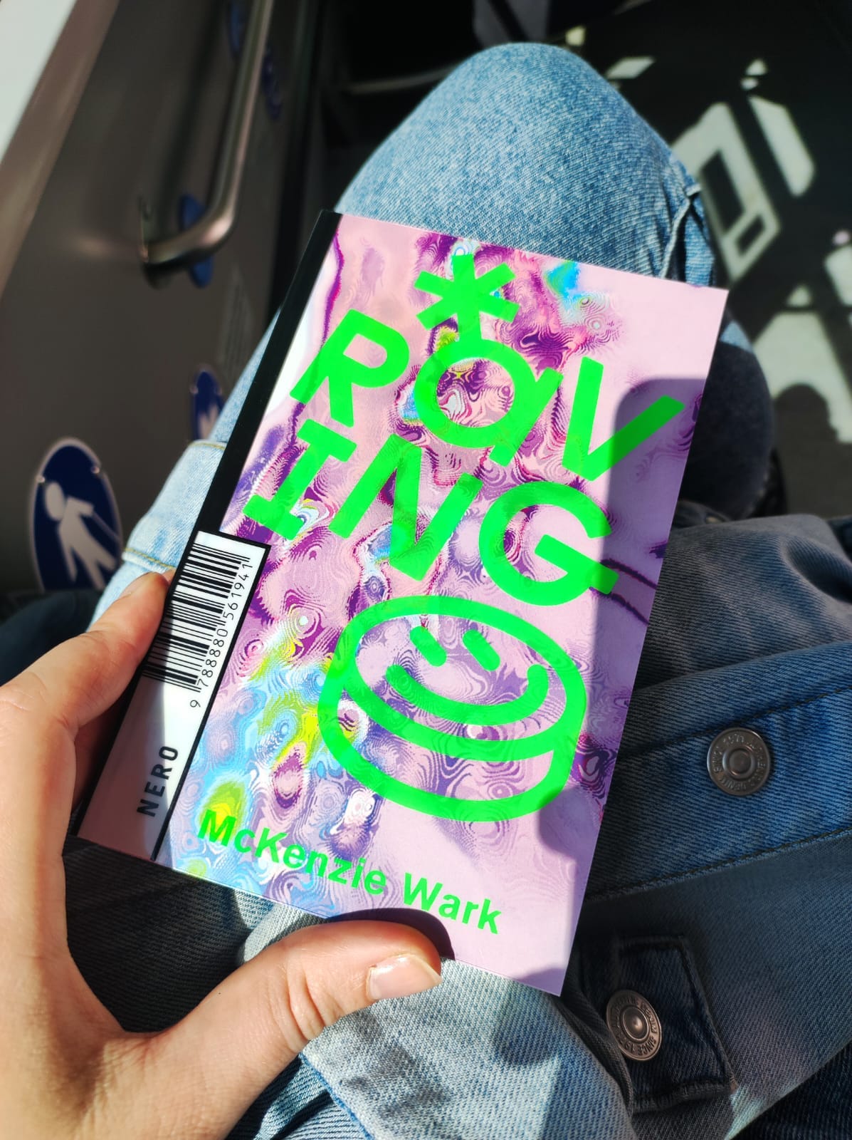 Libro con copertina con un motivo verde e lilla. Il libro, tenuto con una mano, è appoggiato su delle gambe con dei jeans. Sullo sfondo si intravede un autobus