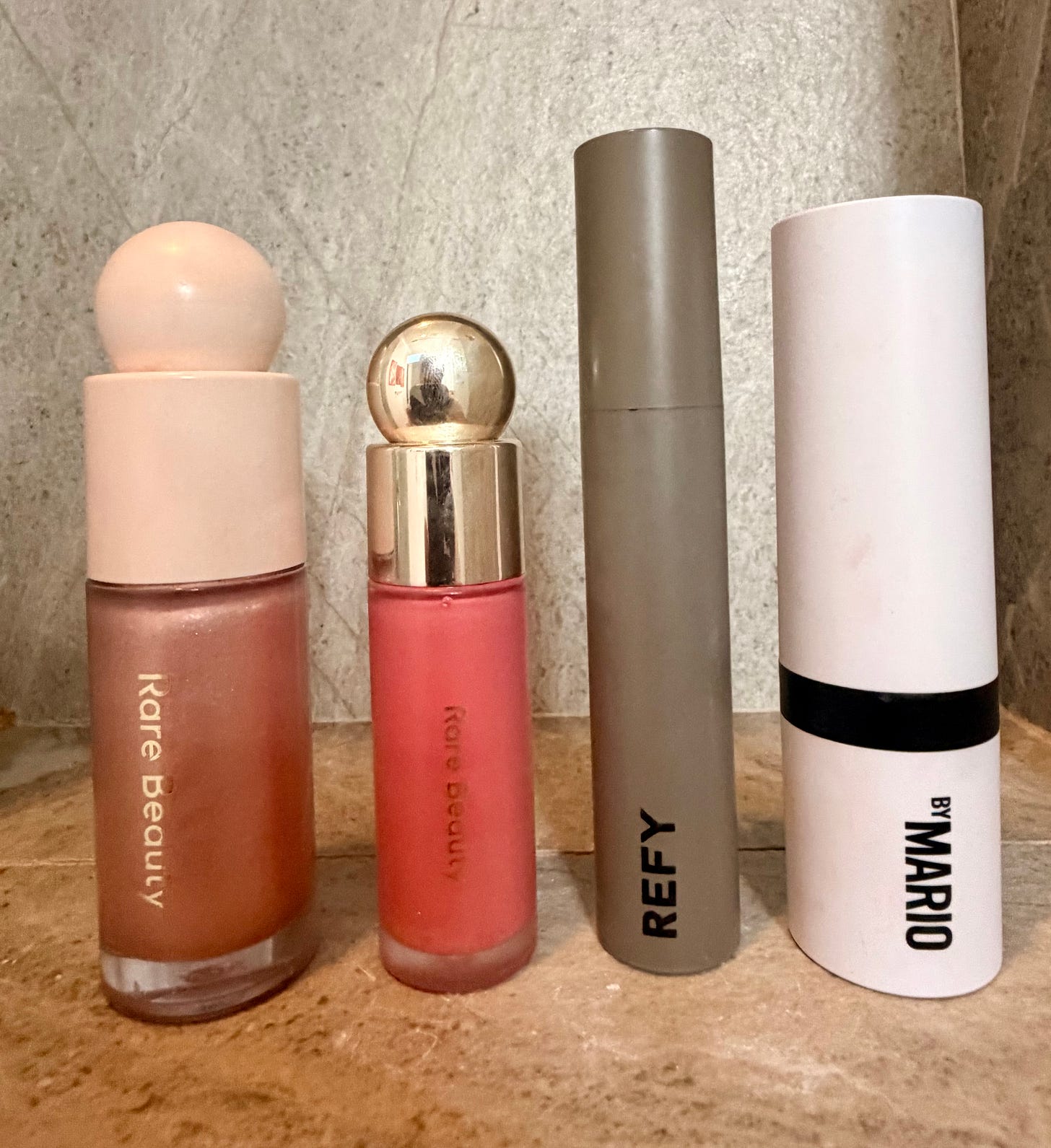 A medium shot photograph of four beauty products standing in a row on a light marble surface against a stone background. From left to right: a tall glass bottle of Rare Beauty Positive Light Liquid Luminizer, a smaller glass bottle of Rare Beauty Soft Pinch Liquid Blush (both with light pink orb caps), a minimalist taupe tube of REFY Brow Sculpt, and a sleek white and black tube of Makeup By Mario MoistureGlow Plumping Lip Serum. The specific product names and brands are clearly visible. A medium shot photograph of four beauty products standing in a row on a light marble surface against a stone background. From left to right: a tall glass bottle of Rare Beauty Positive Light Liquid Luminizer, a smaller glass bottle of Rare Beauty Soft Pinch Liquid Blush (both with light pink orb caps), a minimalist taupe tube of REFY Brow Sculpt, and a sleek white and black tube of Makeup By Mario MoistureGlow Plumping Lip Serum. The specific product names and brands are clearly visible.