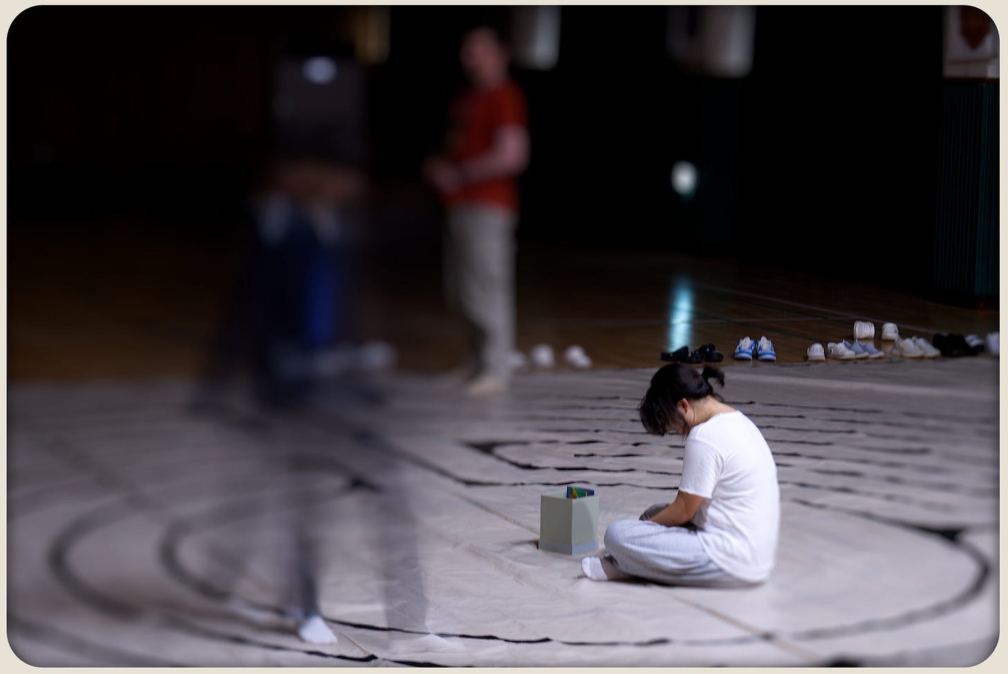 Student sitting at the center of a taped labyrinth on a tarp beside a small box of cards; blurred figures move around in the dim auditorium.