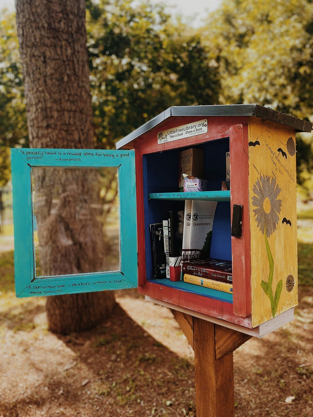 A little library shelf near a tree. It’s painted in blue, red and yellow. There are some books inside.